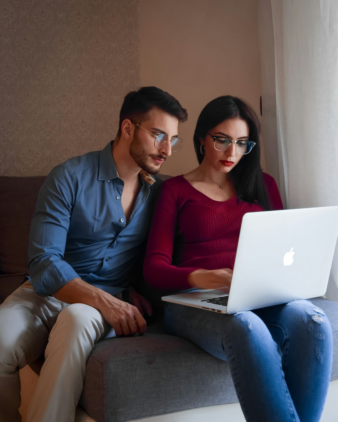 A couple sits closely together on a couch, both intently focused on a laptop. The man, wearing a blue shirt and glasses, leans in, while the woman, in a burgundy top and glasses, types on the laptop. The soft lighting and intimate setting highlight their collaborative effort and modern style.