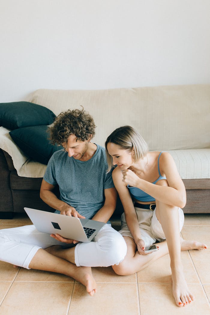 A couple sits barefoot on the floor at home, sharing a laptop for online shopping.