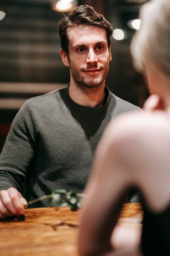 Romantic man in casual clothes with flower in hand sitting at wooden table with unrecognizable girlfriend during date in cafe
